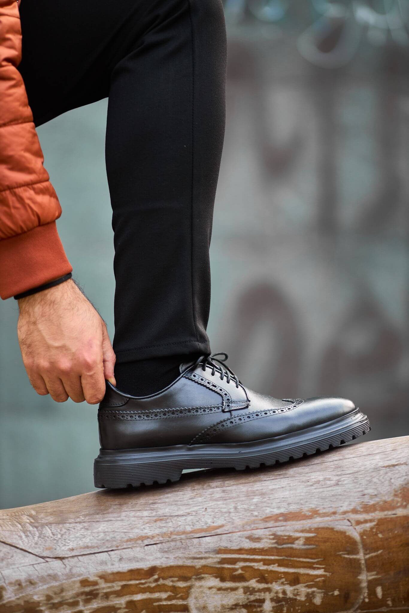 man wearing black leather wingtip brogue derby shoes while sitting on a wooden bench, showcasing detailed perforations and a chunky sole.