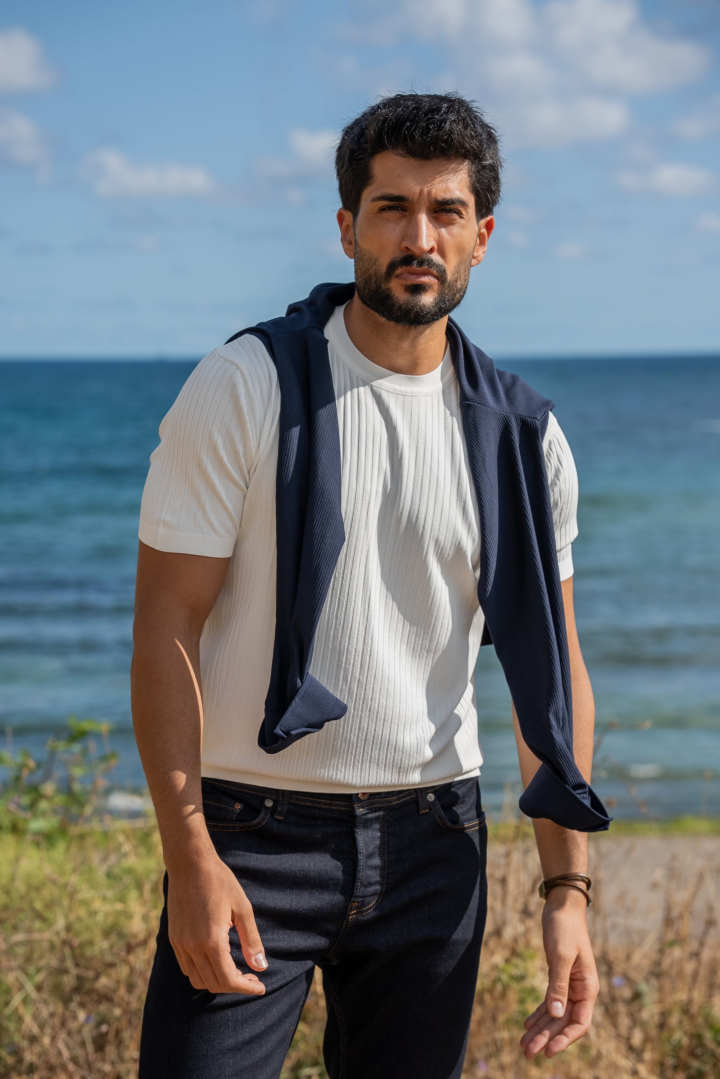 A man wearing a white ribbed knitted t-shirt with a navy sweater draped over his shoulders, standing by the sea under a sunny blue sky.