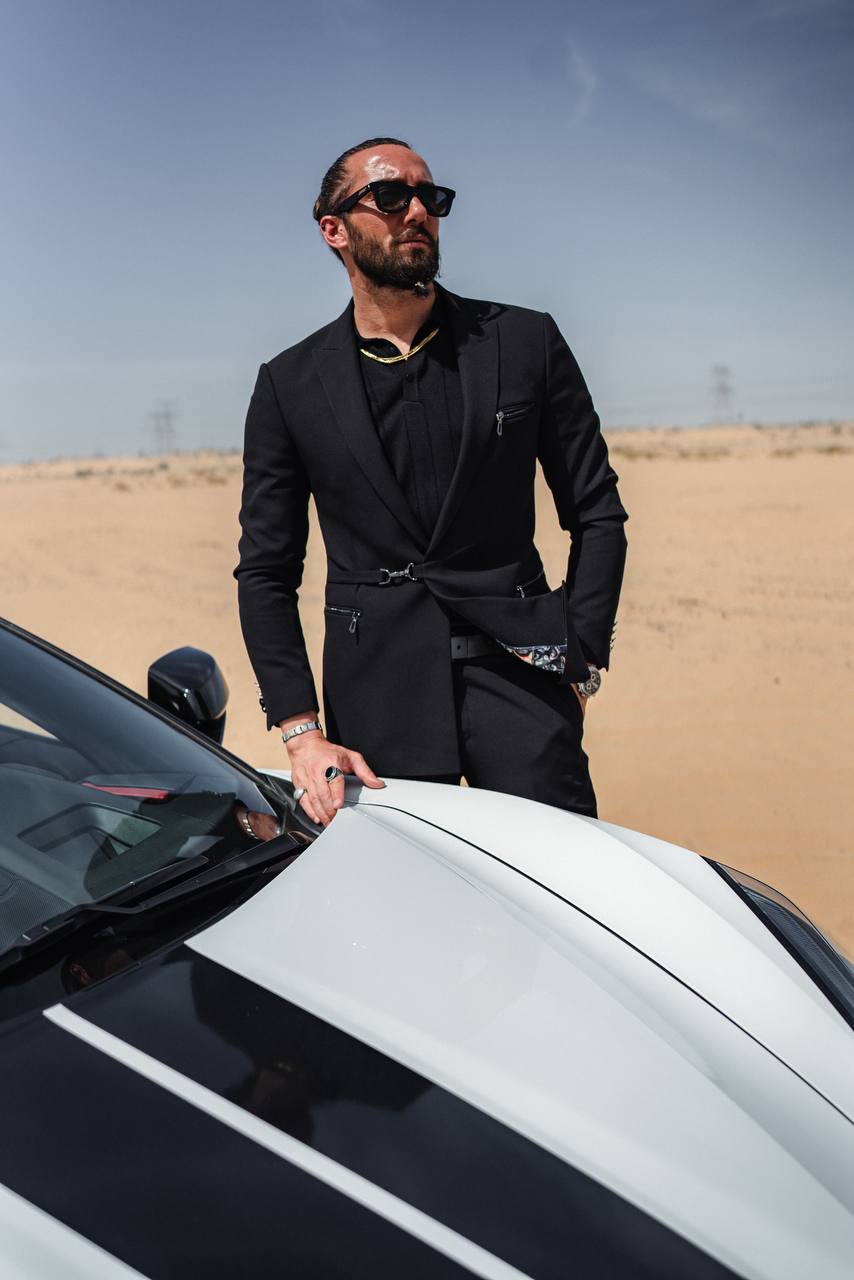 Man in slim-fit black HolloMen summer suit with zippered accents, styled with black shirt and sunglasses against a desert luxury car backdrop