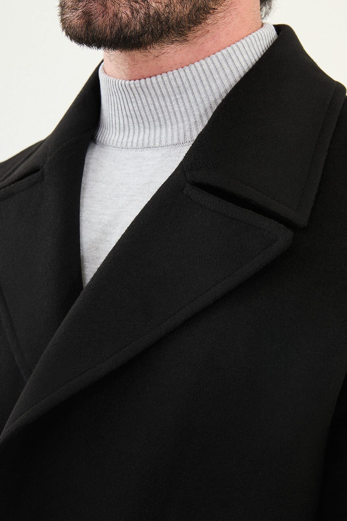 Man wearing a black slim fit double-breasted long coat with wide collar and button closure, styled over a light gray turtleneck.