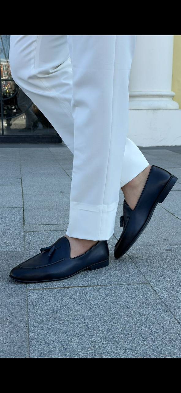 Men’s black leather tassel loafers paired with white tailored trousers, showcasing a sleek slip-on design and classic tassel detail.