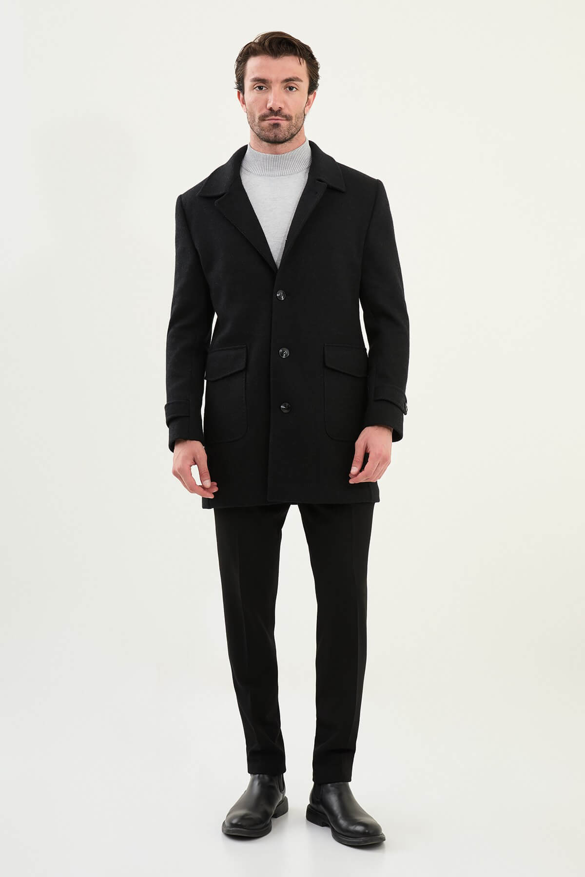A man wearing a slim fit black single-breasted coat over a light grey turtleneck, styled with black trousers and black leather boots.