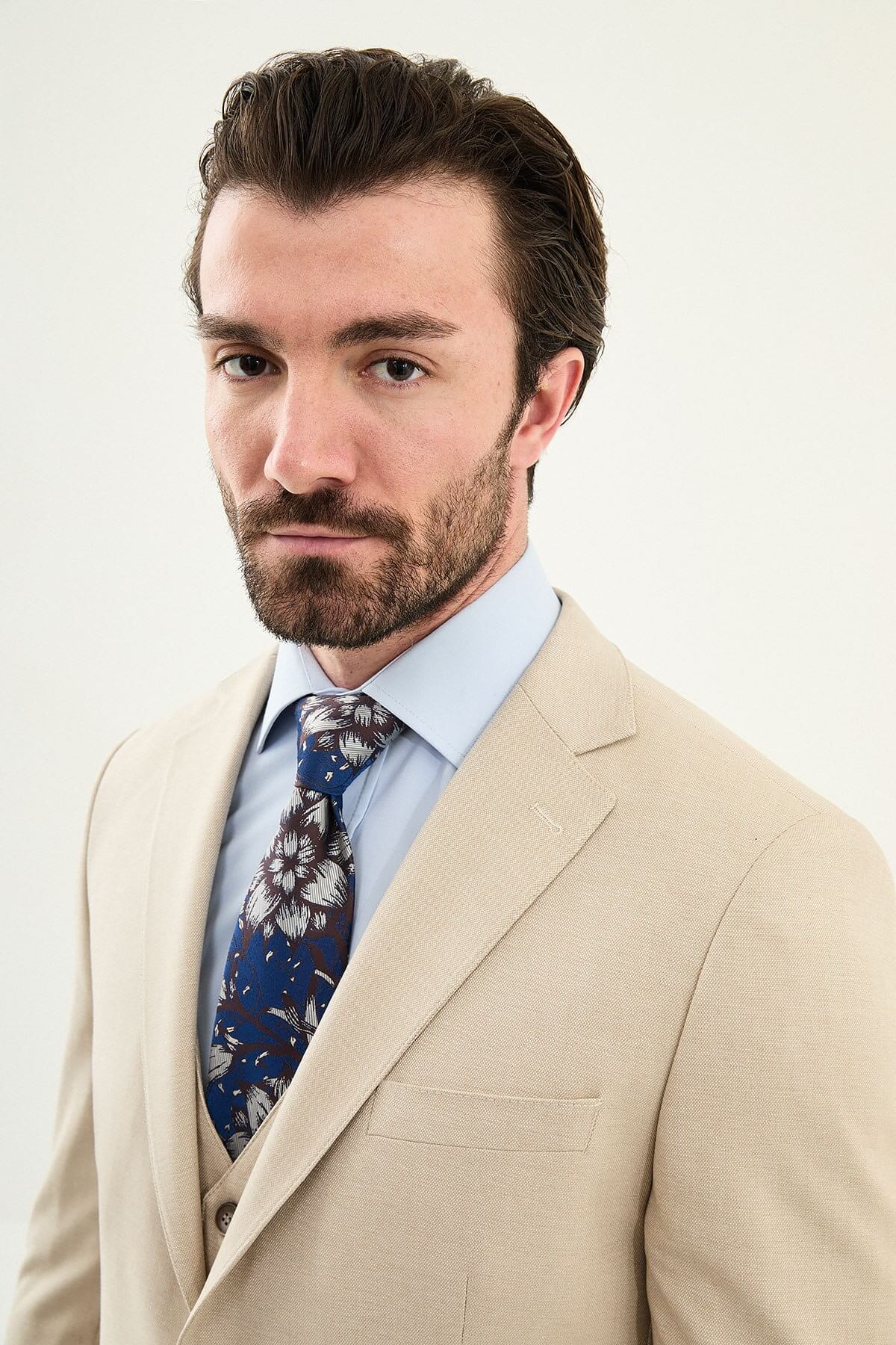 Model wearing a beige slim-fit cotton-linen three-piece suit with a pale blue dress shirt, navy floral tie, and matching tailored trousers.