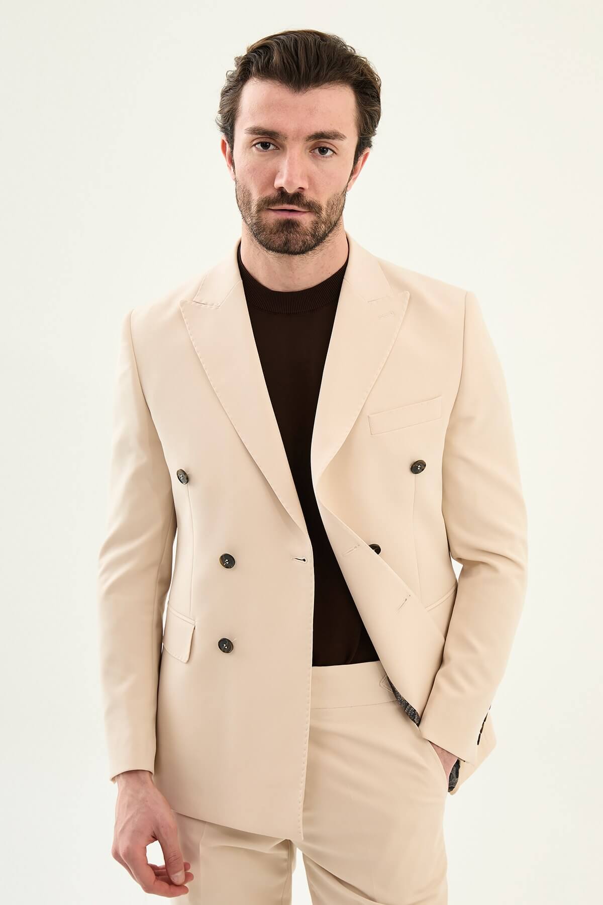 Model wearing a beige slim-fit double-breasted suit with peak lapels, dark buttons, matching trousers, a dark brown knit top, and brown leather dress shoes.