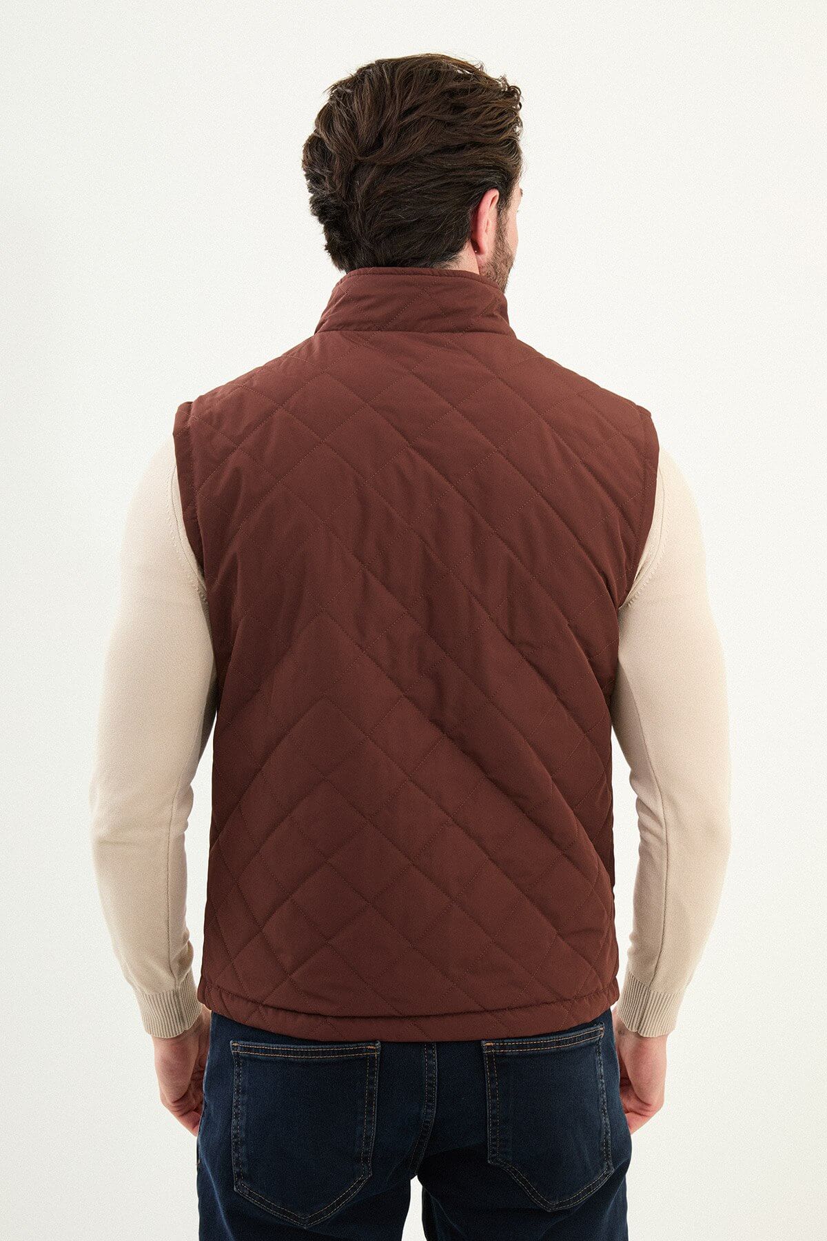 Tile rust diamond-quilted men’s technical vest in matte polyester, styled over a beige turtleneck with dark denim.