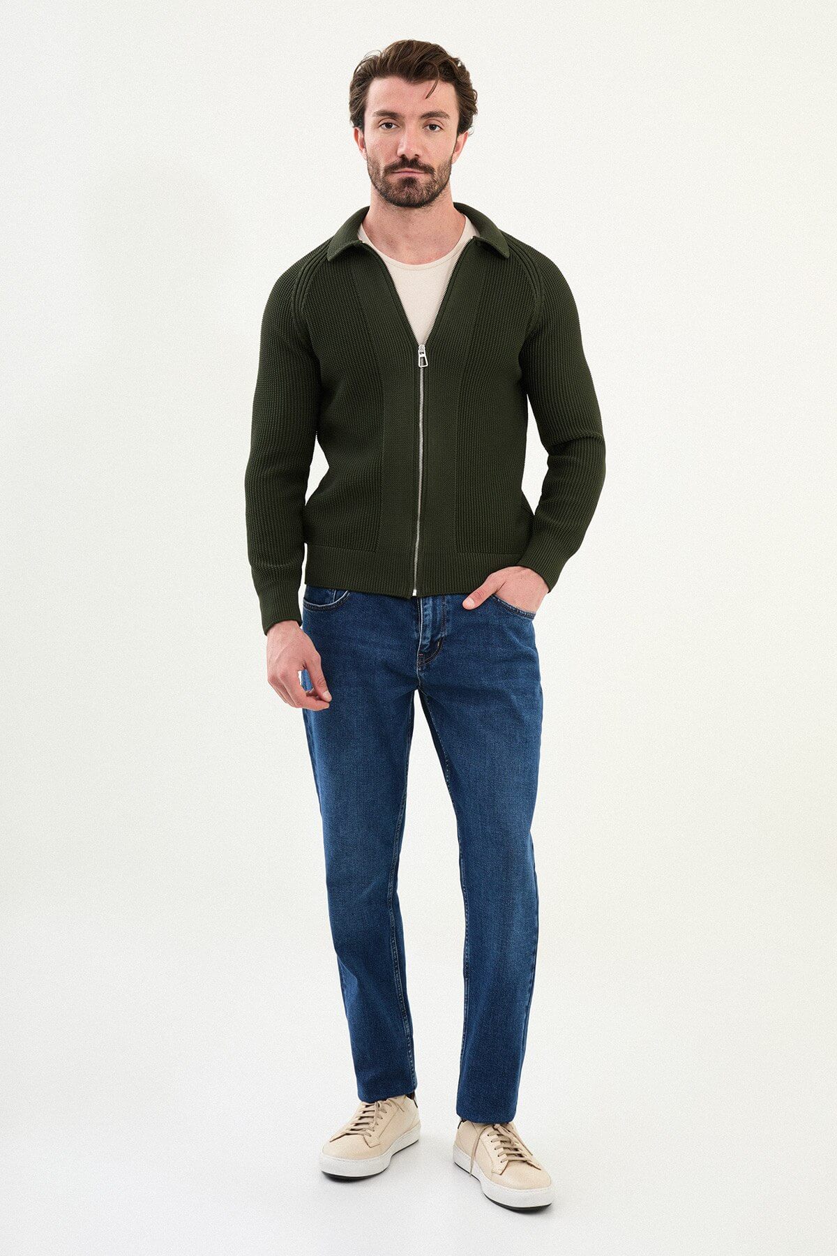 A man wearing a slim fit green ribbed zip-up knitwear cardigan with a collared design, paired with blue jeans and beige sneakers.