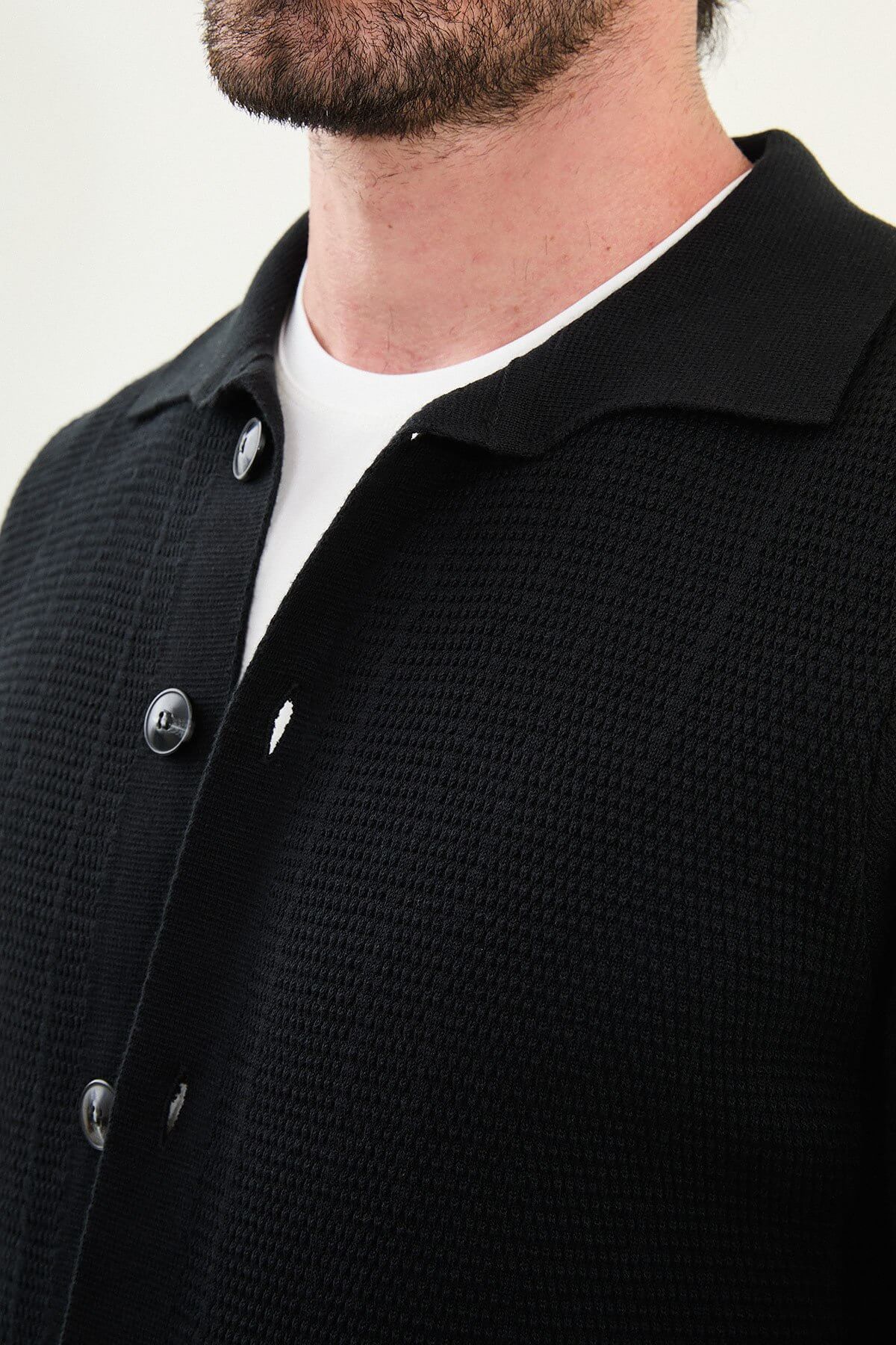 A man wearing a slim fit black button-up cardigan with front pockets, styled with a white T-shirt and grey jeans.