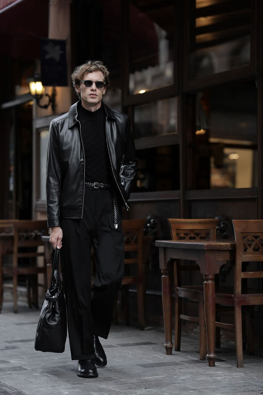 Men's black leather-look jacket styled with a cream knit sweater and black trousers.