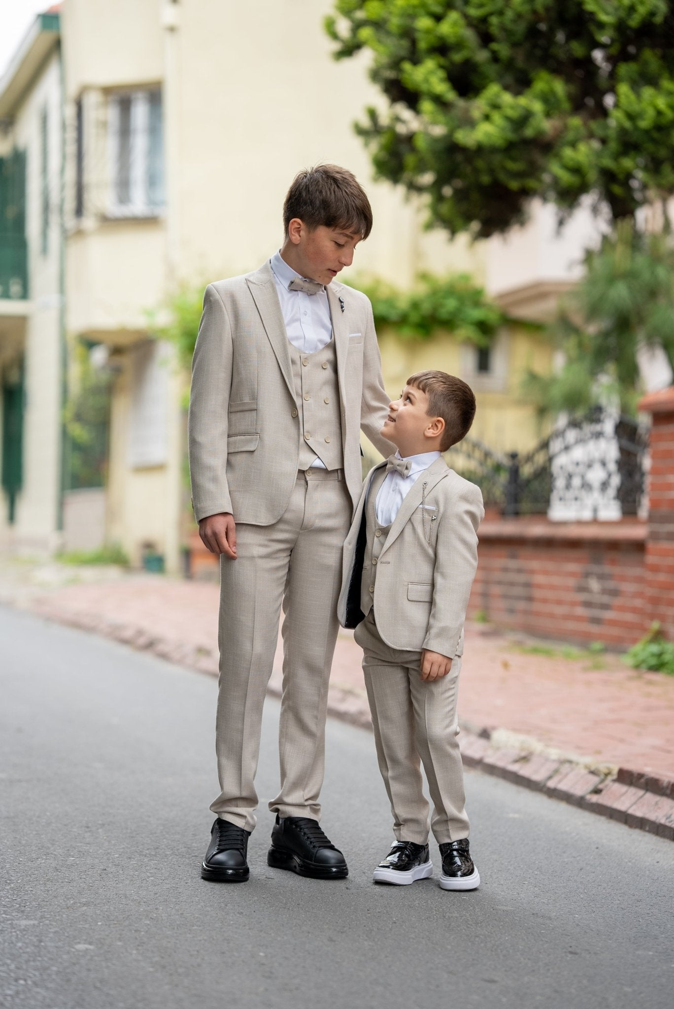 Father and his two sons walking down a street in matching beige tuxedos with elegant bow ties, exuding confidence and style during a formal occasion.