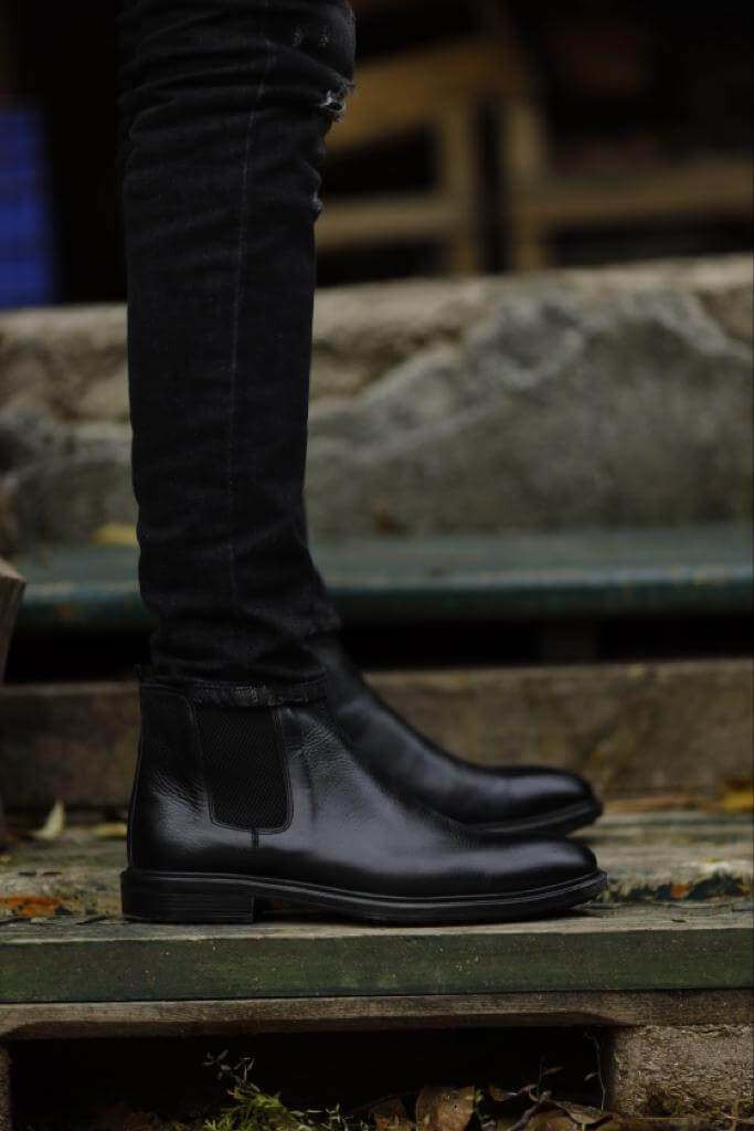 Close-up of a man wearing sleek black Chelsea boots paired with black jeans, standing on rustic wooden logs in an outdoor setting.