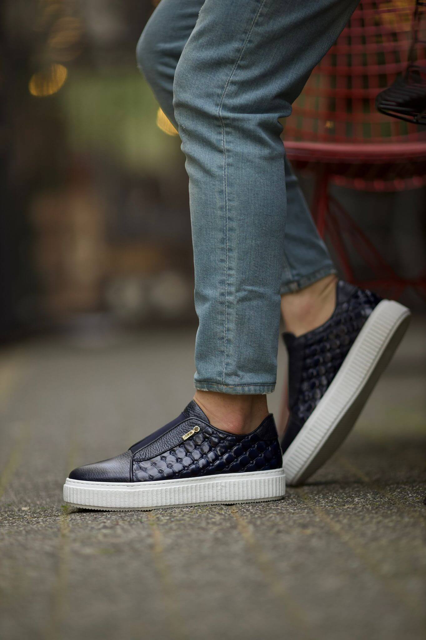 A close-up of a man wearing Seville Quilted Black Slip-On Sneakers, featuring a black leather upper with a quilted side panel, white EVA sole, and subtle side zip detail. Styled with light wash denim.