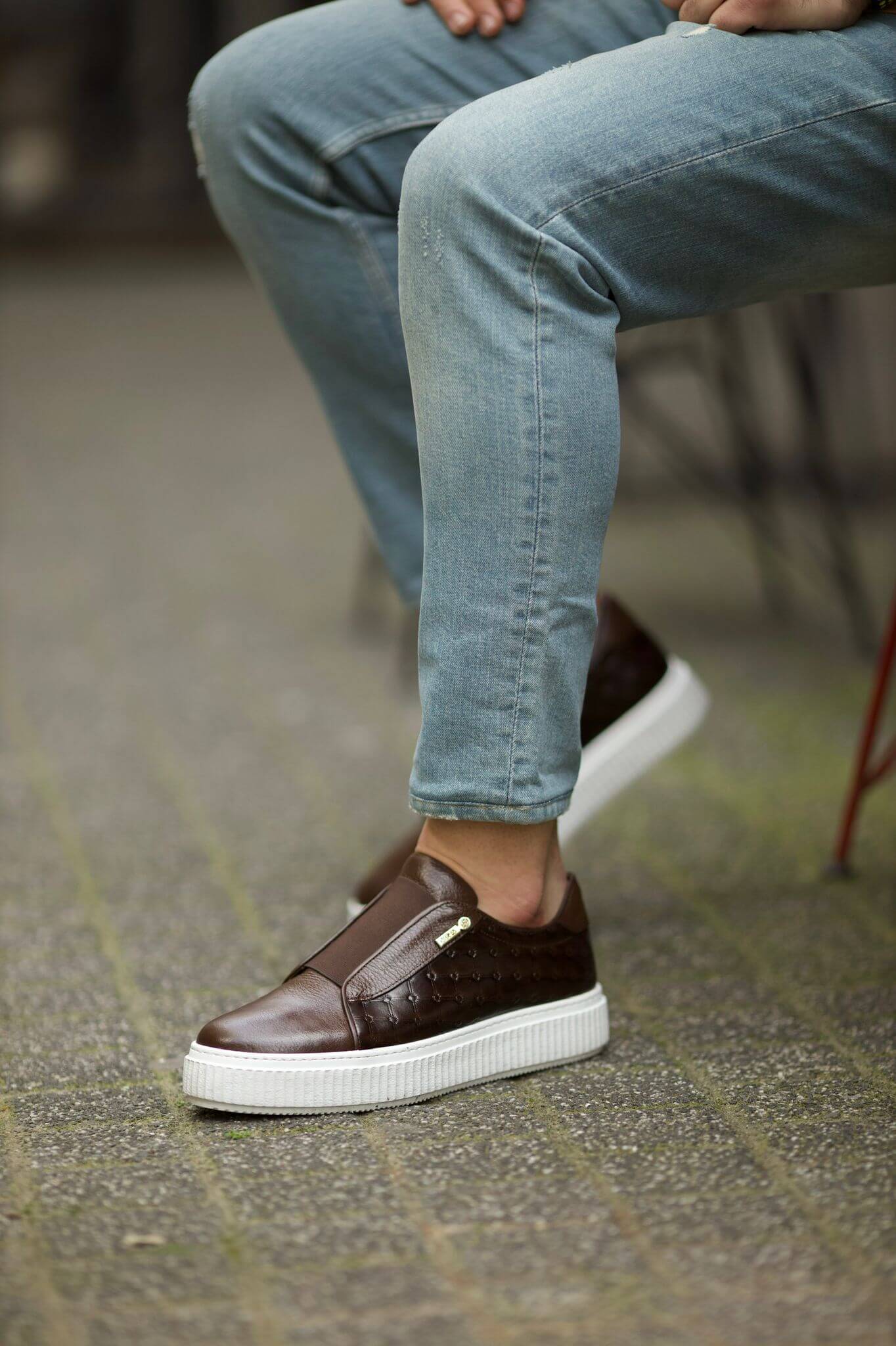 Close-up of a man wearing brown leather slip-on sneakers with quilted sides and white EVA soles, styled with cuffed light blue jeans.