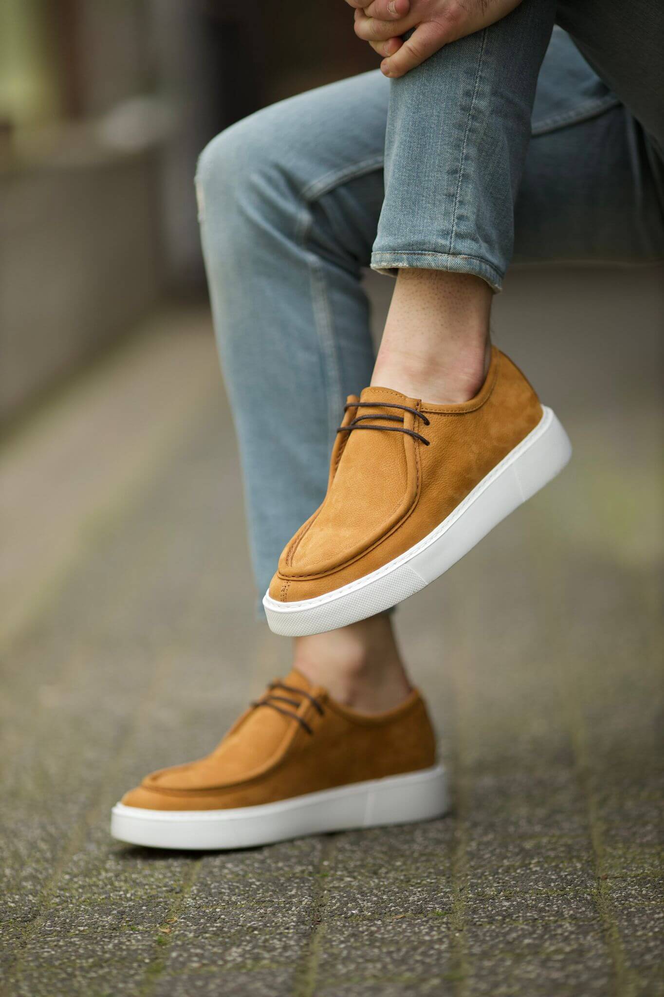 A pair of camel-colored leather derby shoes with black laces and white EVA soles, styled with light blue jeans in an urban outdoor setting.