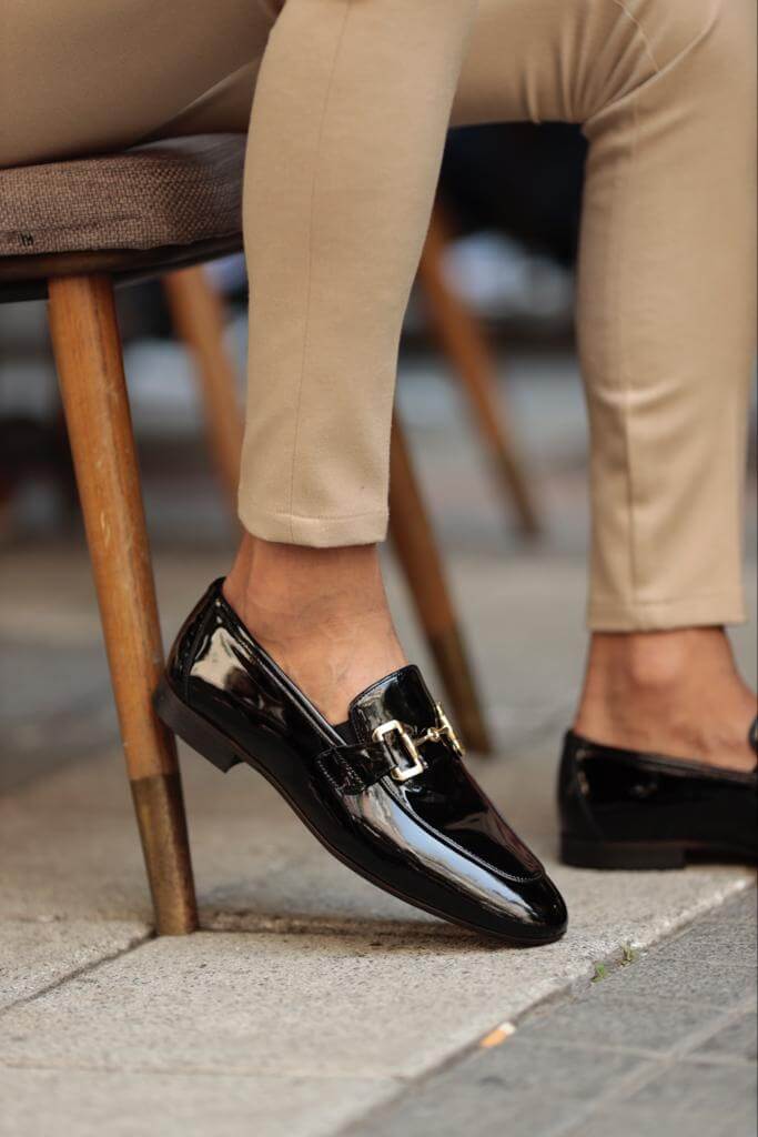 A sleek pair of men's black patent leather loafers with a modern buckle detail, photographed under soft light to highlight the rich shine and sharp design.