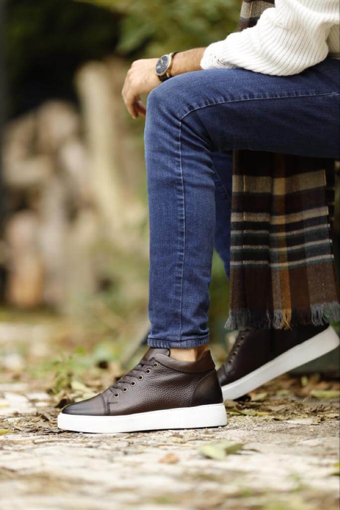 Man wearing high-top brown leather sneakers with white rubber soles and side zippers, styled with fitted blue denim jeans on an urban street setting.