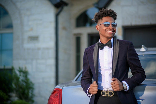A young man posing in a dark navy textured floral tuxedo jacket with black satin shawl lapels, a black bow tie, and a gold Ferragamo belt buckle, leaning against a silver luxury car for a modern prom look