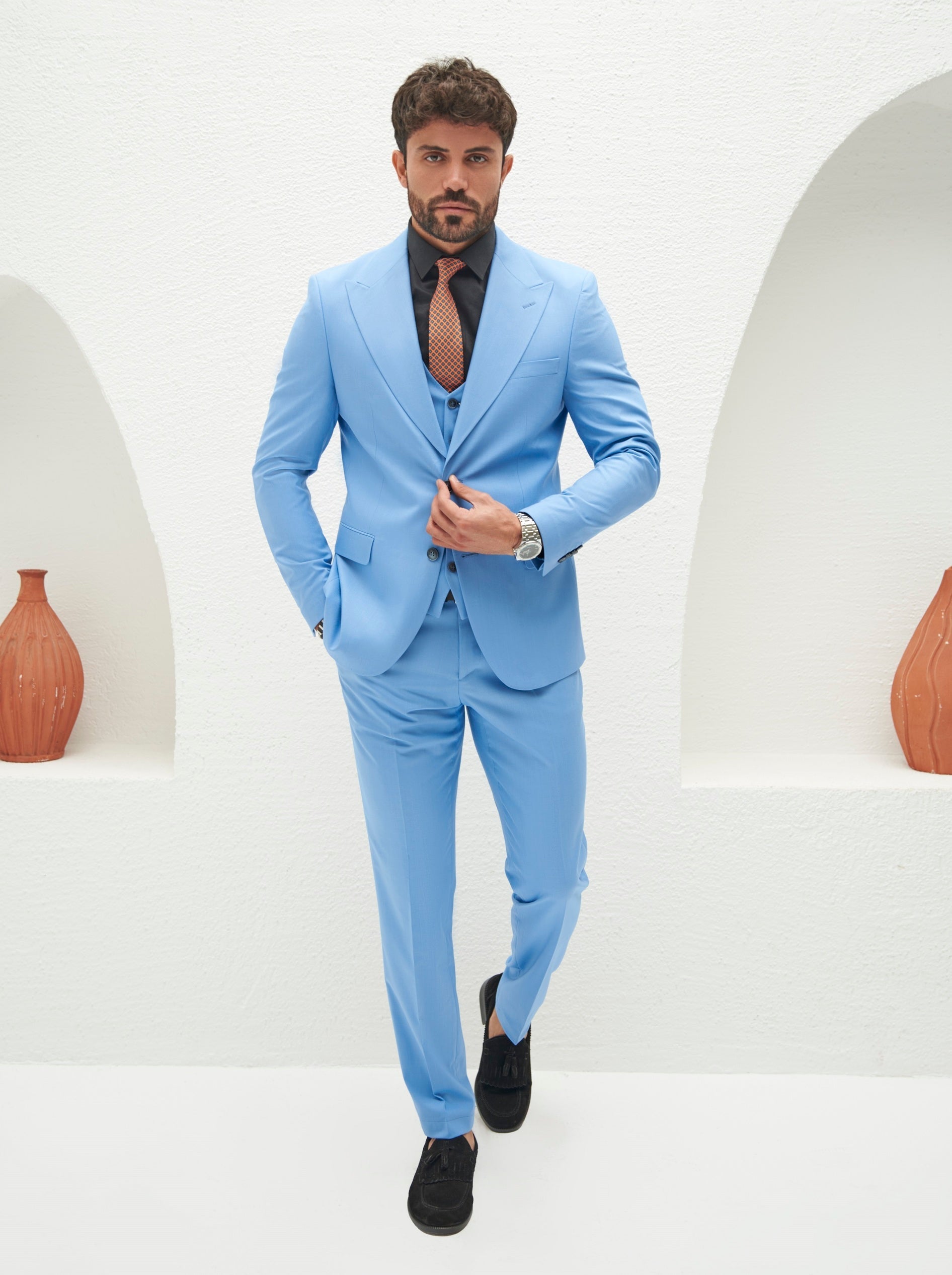 Men’s sky-blue slim-fit 3-piece suit with blazer, waistcoat, and trousers, styled with a white shirt and patterned tie in a modern interior setting.