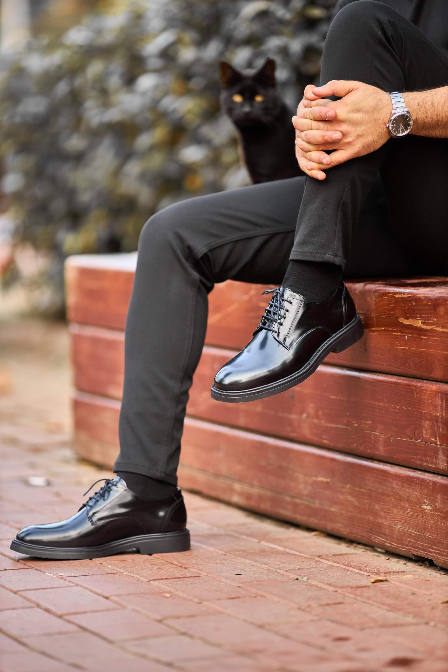 Black polished leather Derby shoes worn with black trousers, styled on outdoor wooden bench.