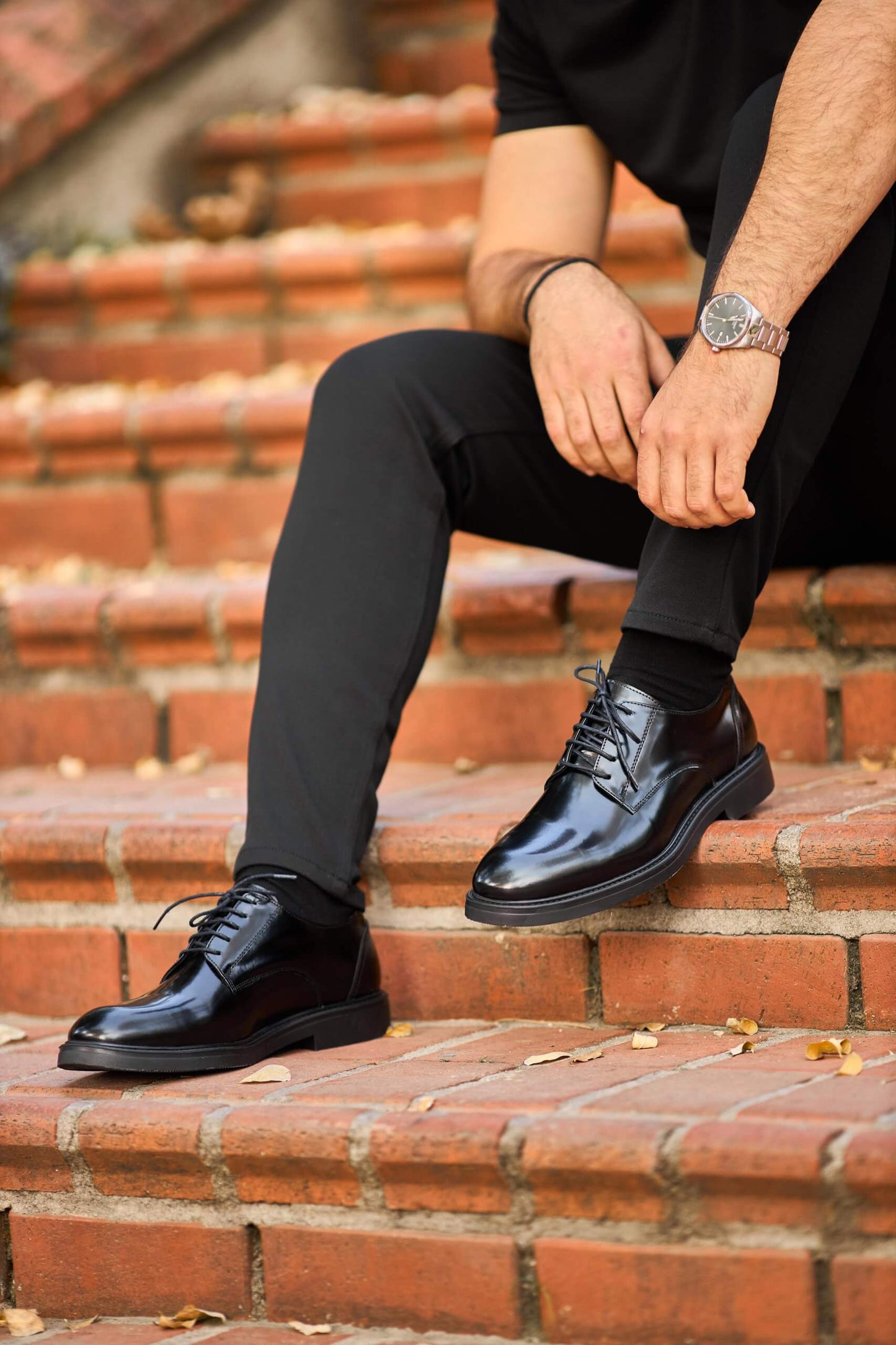 Black polished leather Derby shoes worn with black trousers, styled on outdoor wooden bench.
