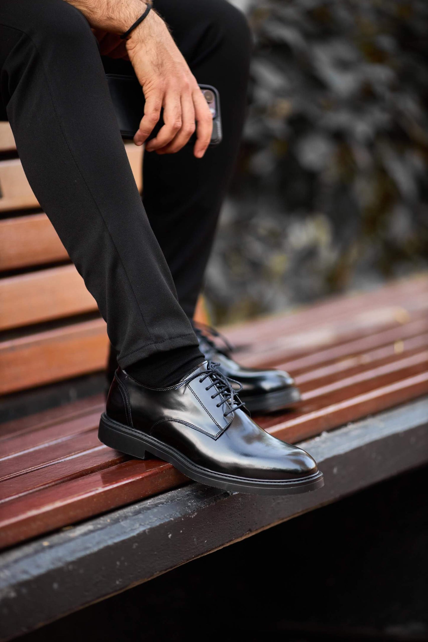 Black polished leather Derby shoes worn with black trousers, styled on outdoor wooden bench.
