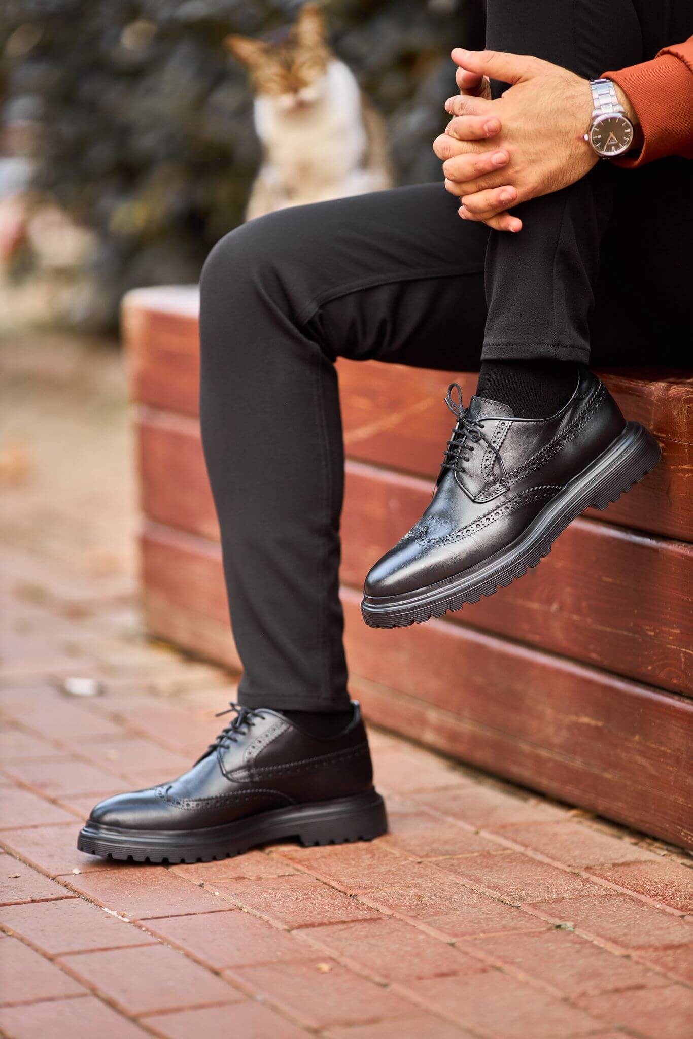 man wearing black leather wingtip brogue derby shoes while sitting on a wooden bench, showcasing detailed perforations and a chunky sole.