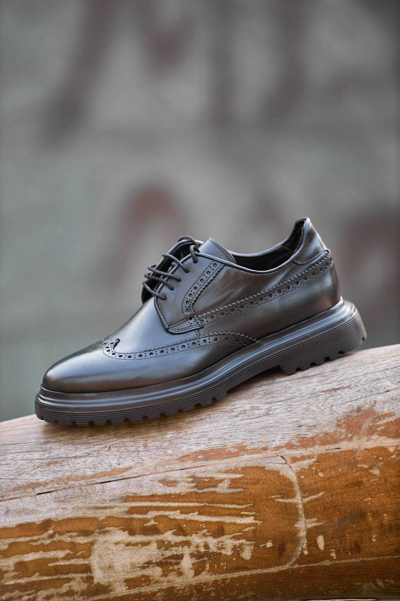 man wearing black leather wingtip brogue derby shoes while sitting on a wooden bench, showcasing detailed perforations and a chunky sole.