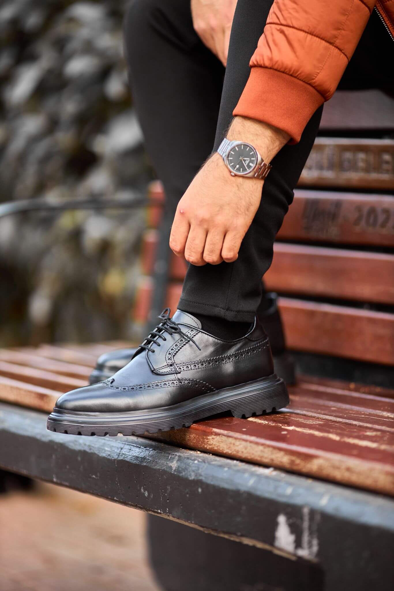 man wearing black leather wingtip brogue derby shoes while sitting on a wooden bench, showcasing detailed perforations and a chunky sole.
