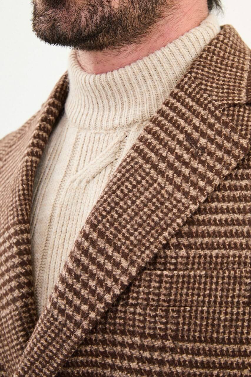 Men’s brown double-breasted herringbone wool-blend longline overcoat with extra-wide peak lapels, deep overlap front, and styled with a cream turtleneck.