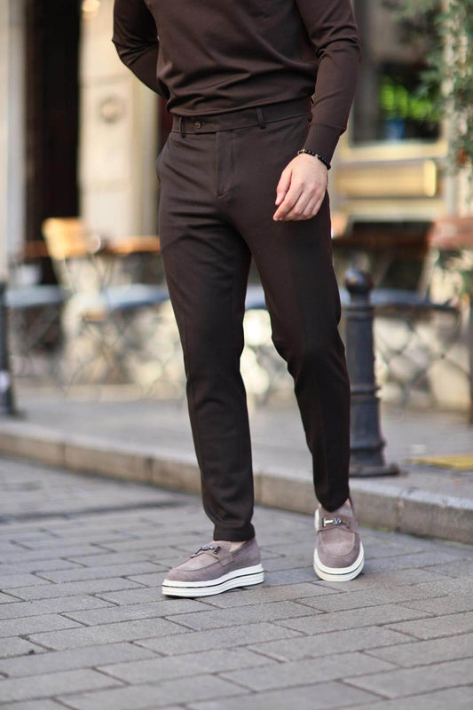 Man wearing brown slim-fit trousers paired with grey loafers, standing casually on a cobblestone street.