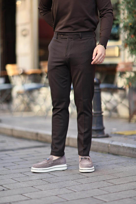 Man wearing brown slim-fit trousers paired with grey loafers, standing casually on a cobblestone street.