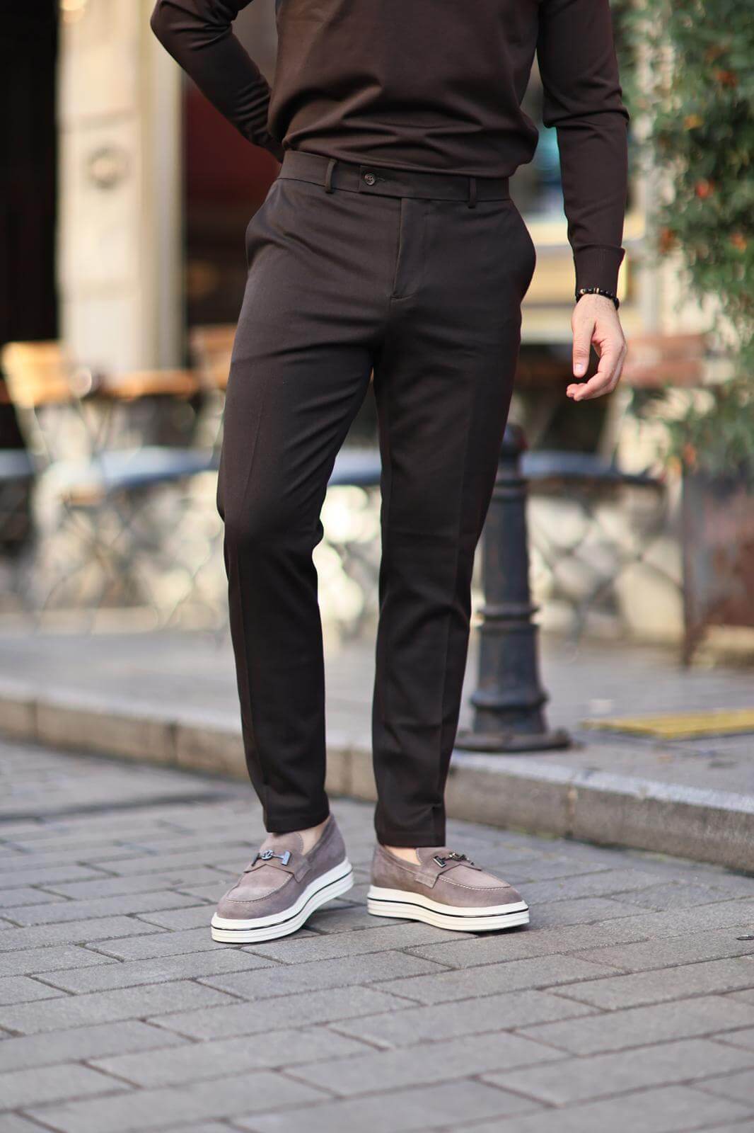 Man wearing brown slim-fit trousers paired with grey loafers, standing casually on a cobblestone street.