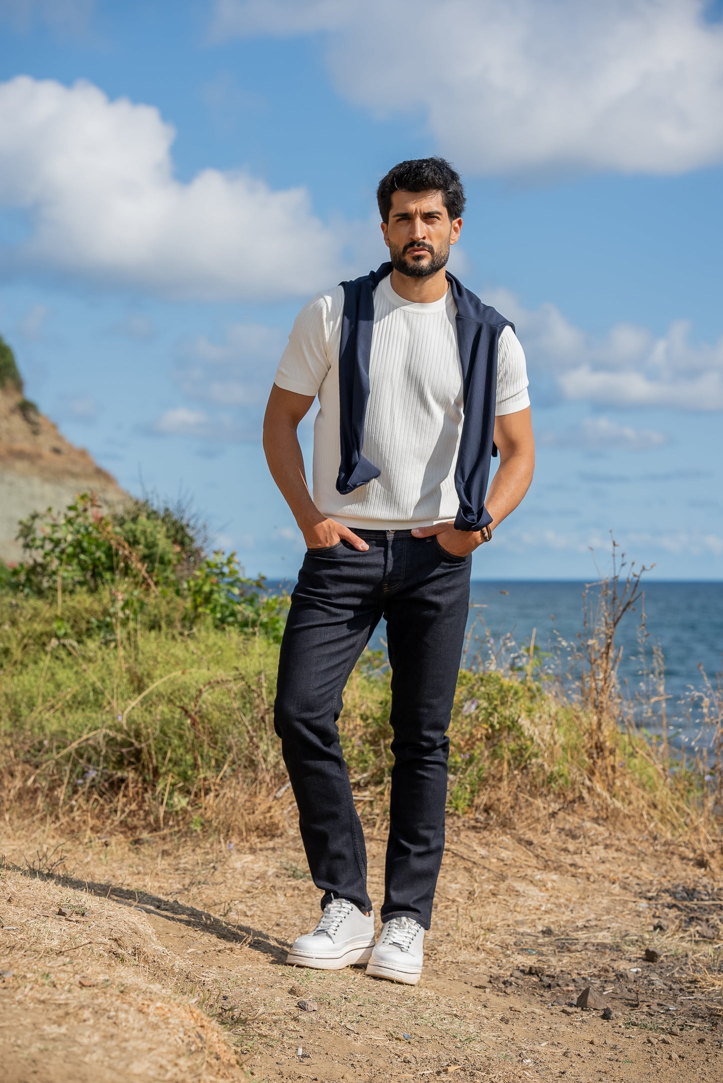 A man wearing a white ribbed knitted t-shirt with a navy sweater draped over his shoulders, standing by the sea under a sunny blue sky.