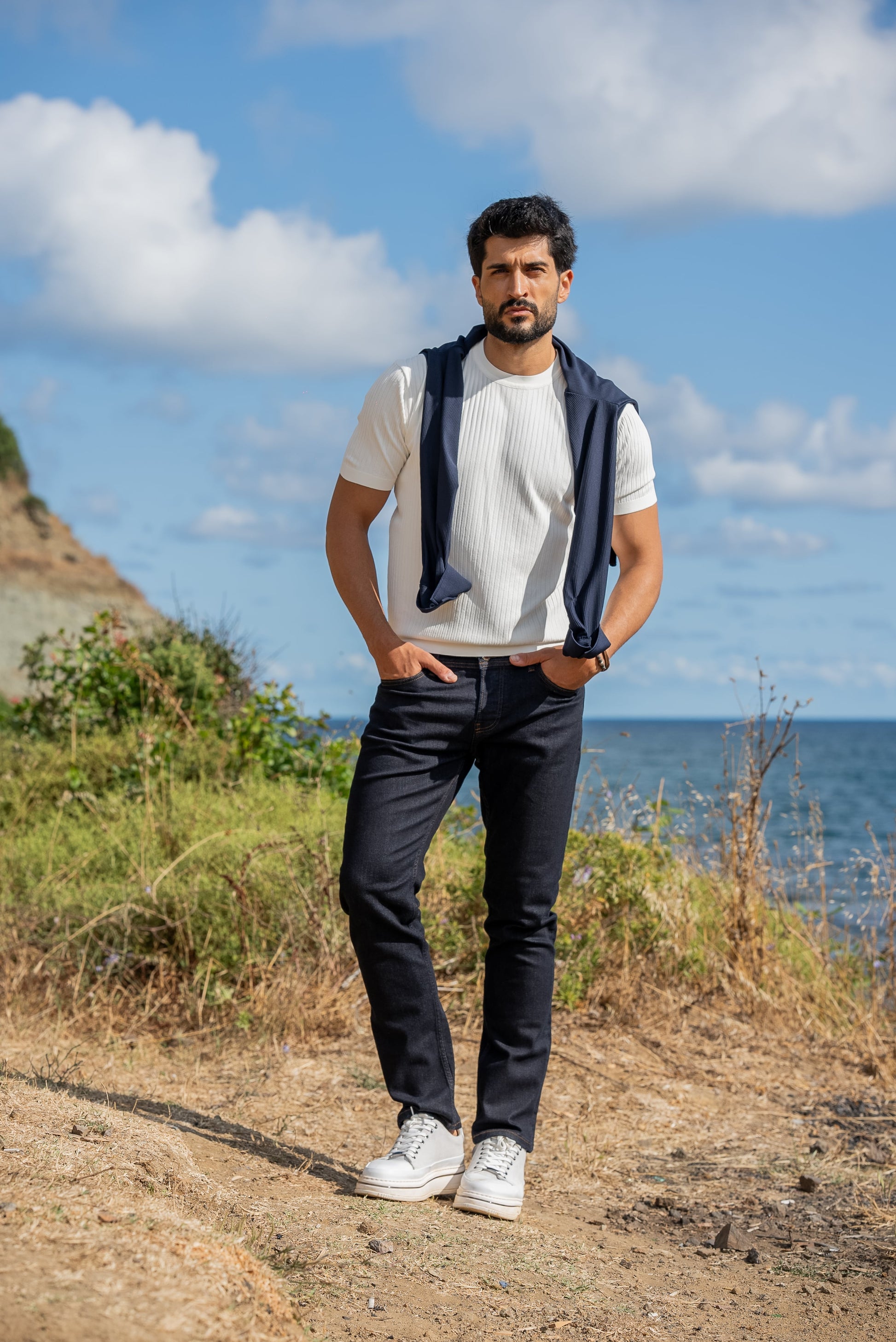 A man wearing a white ribbed knitted t-shirt with a navy sweater draped over his shoulders, standing by the sea under a sunny blue sky.