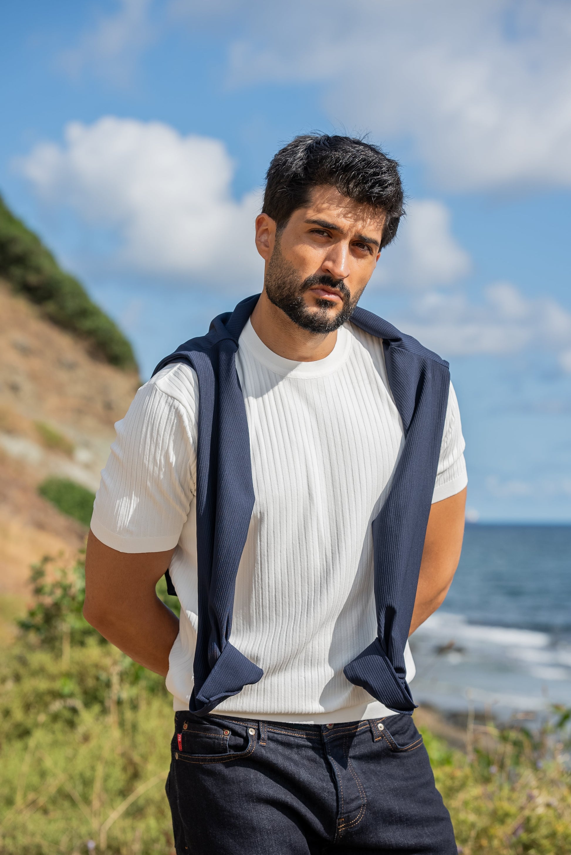 A man wearing a white ribbed knitted t-shirt with a navy sweater draped over his shoulders, standing by the sea under a sunny blue sky.