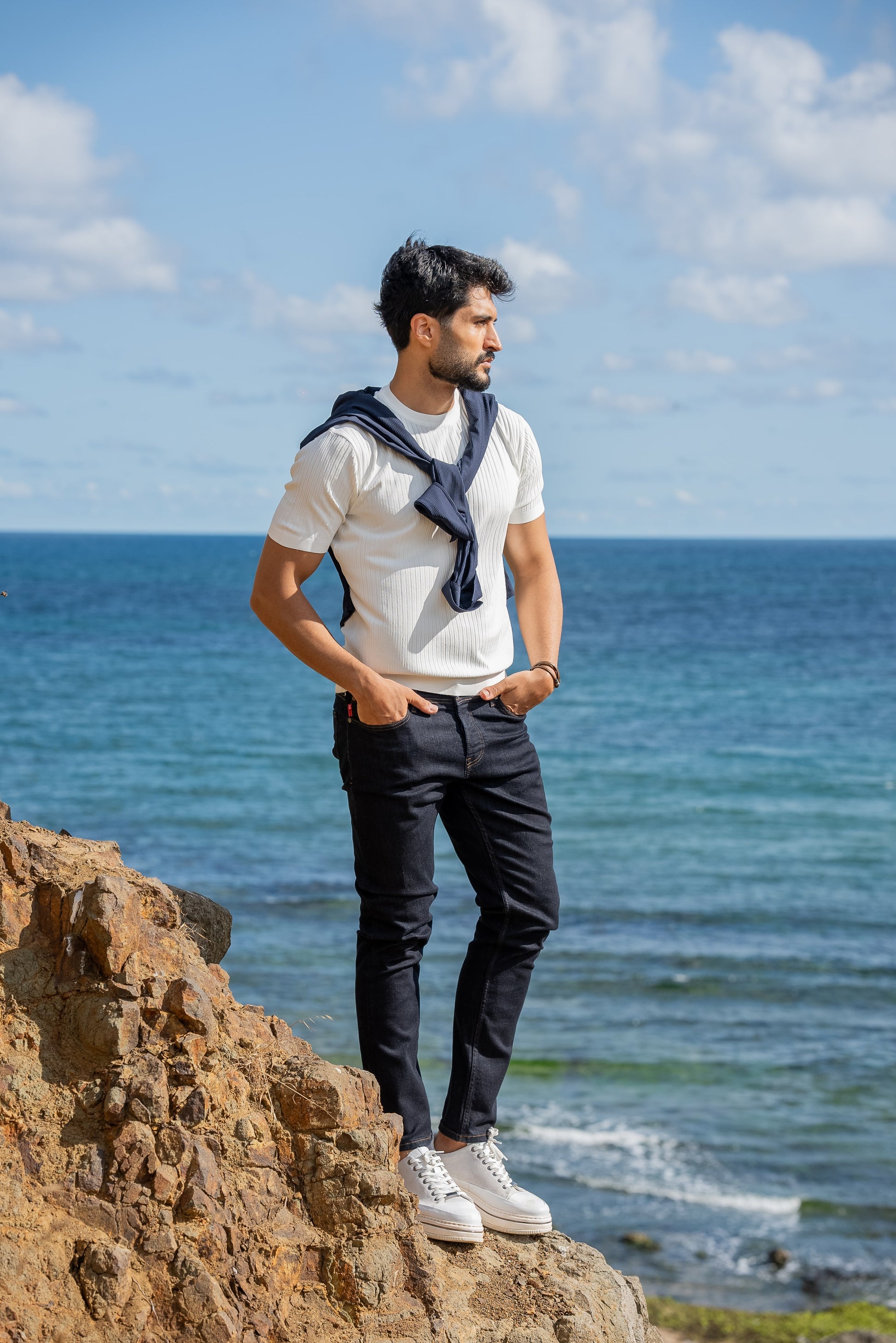 A man wearing a white ribbed knitted t-shirt with a navy sweater draped over his shoulders, standing by the sea under a sunny blue sky.