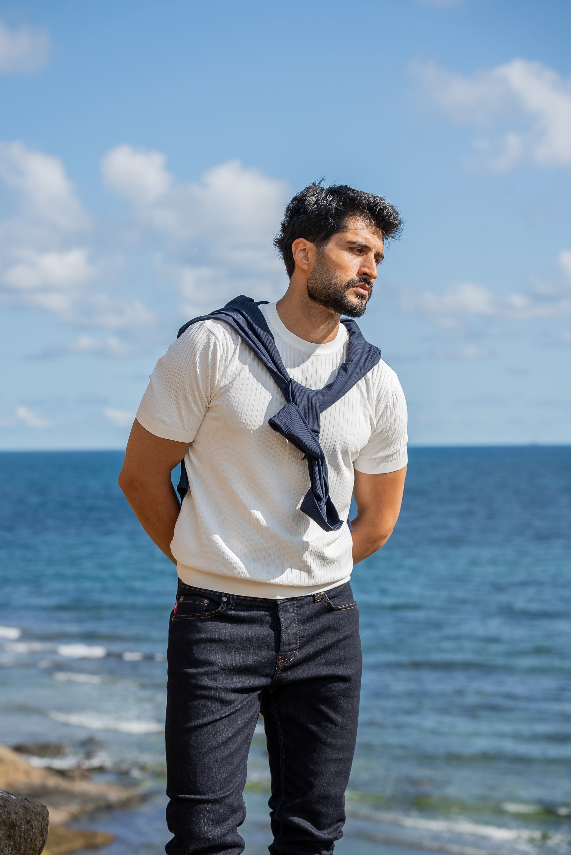 A man wearing a white ribbed knitted t-shirt with a navy sweater draped over his shoulders, standing by the sea under a sunny blue sky.