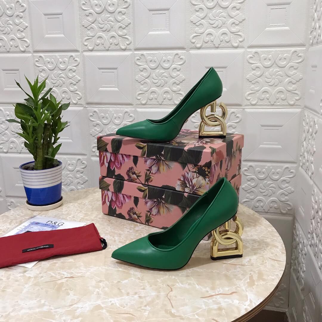 Dolce & Gabbana green leather pumps with a distinctive gold sculptural "DG" logo block heel, displayed on a pink floral patterned box.

