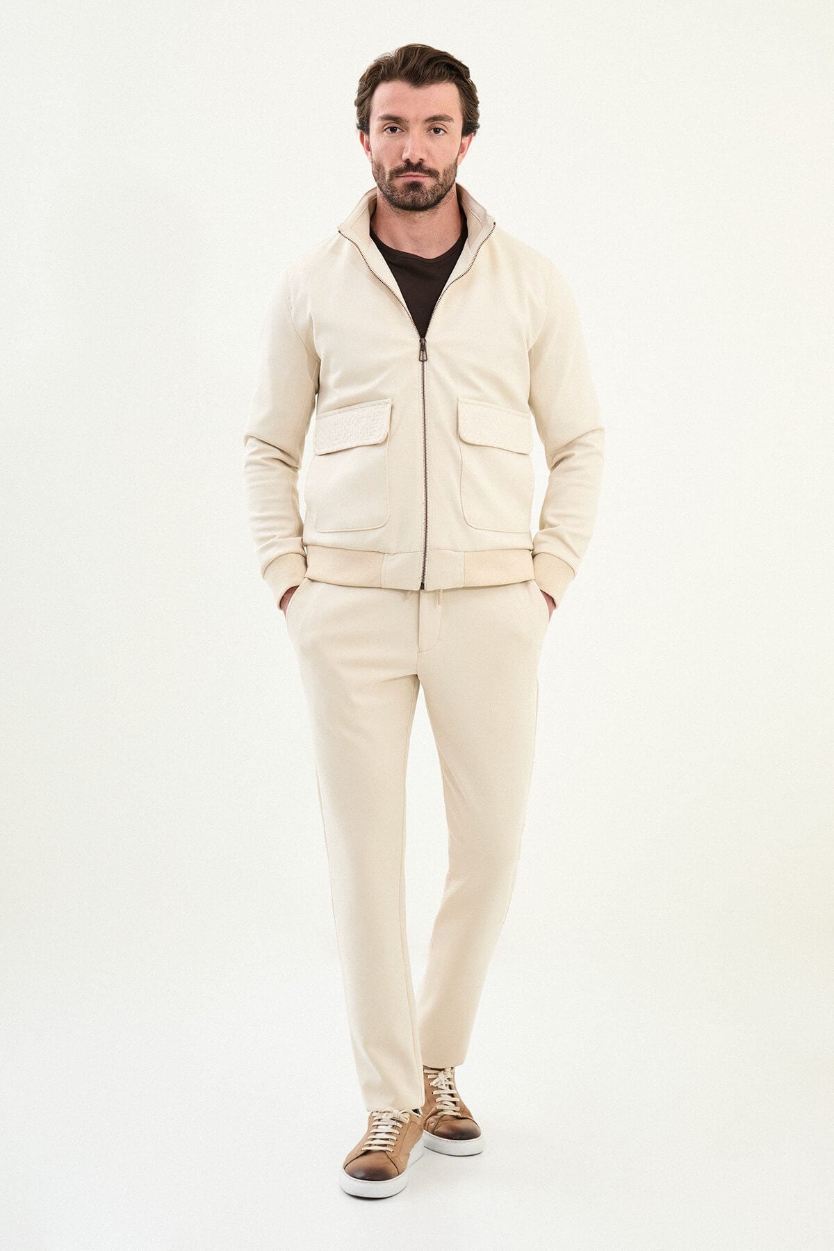 A male model wearing a beige slim fit jacket with zip closure and matching trousers, styled with brown sneakers.