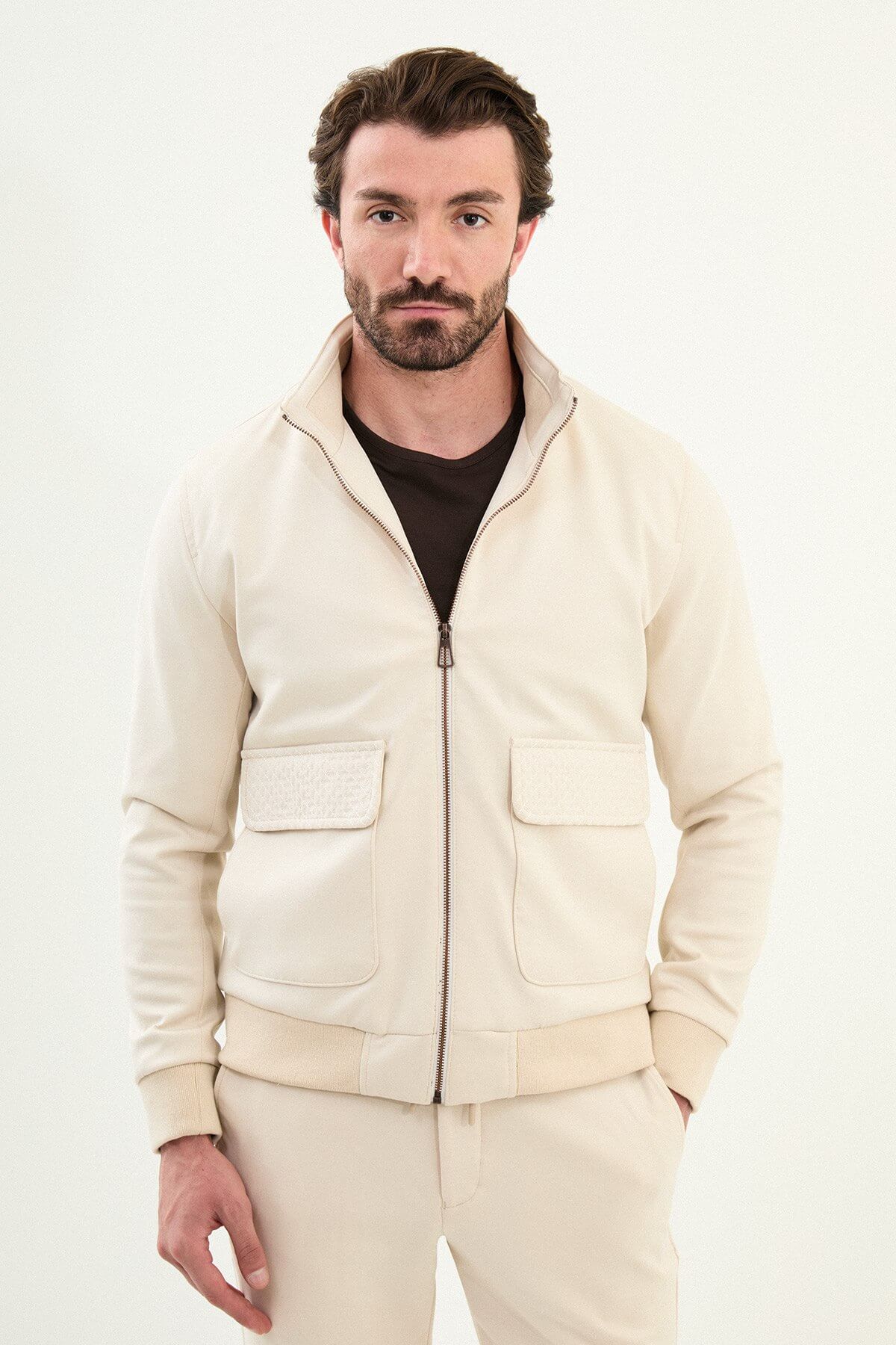 A male model wearing a beige slim fit jacket with zip closure and matching trousers, styled with brown sneakers.