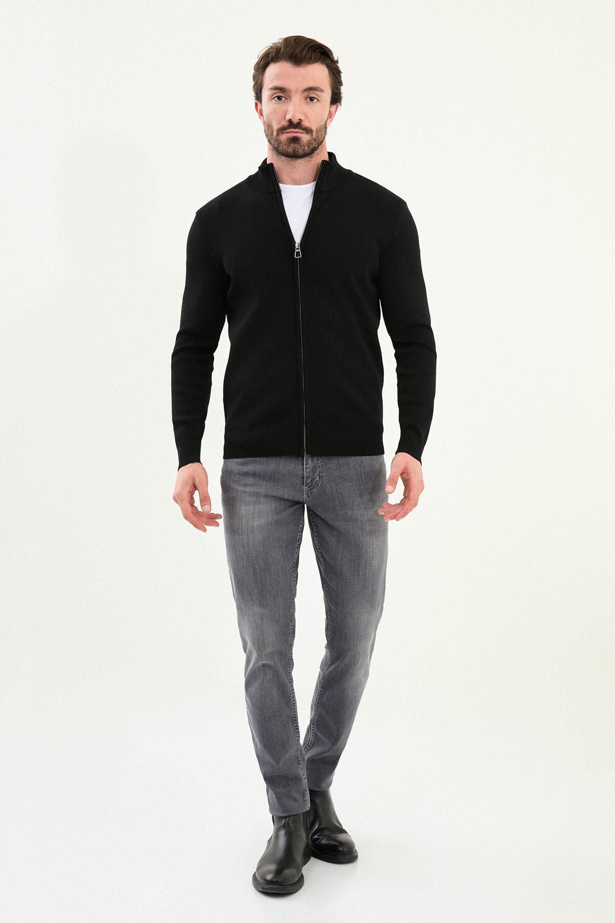 A man wearing a slim fit black zip cardigan over a white T-shirt, styled with grey jeans and black boots.