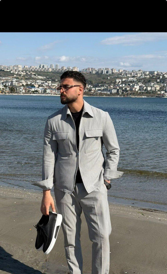 Men’s light grey co-ord set featuring a zip-up jacket with chest pockets and slim-fit trousers, styled in an outdoor waterfront setting.