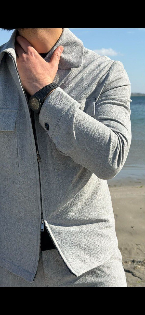 Men’s light grey co-ord set featuring a zip-up jacket with chest pockets and slim-fit trousers, styled in an outdoor waterfront setting.