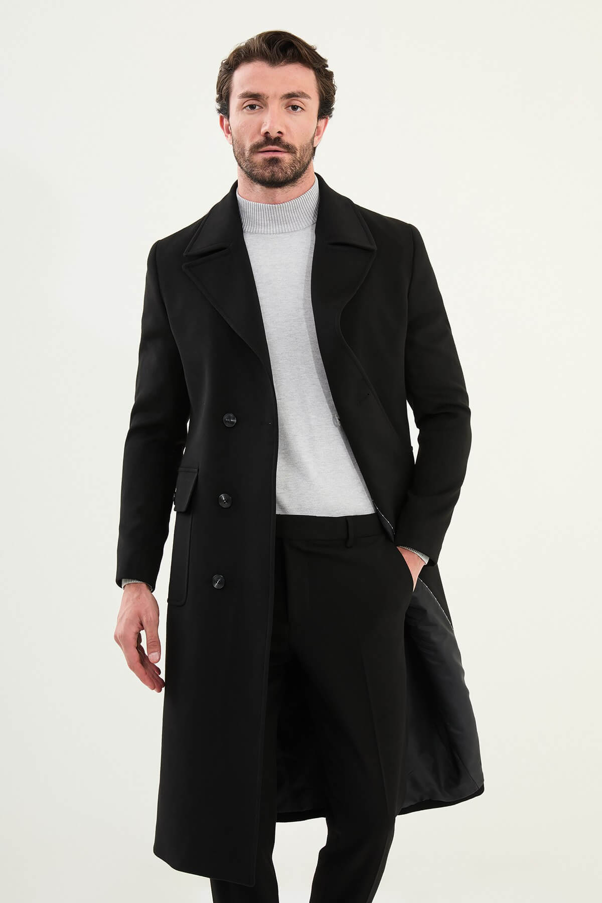 Man wearing a black slim fit double-breasted long coat with wide collar and button closure, styled over a light gray turtleneck.