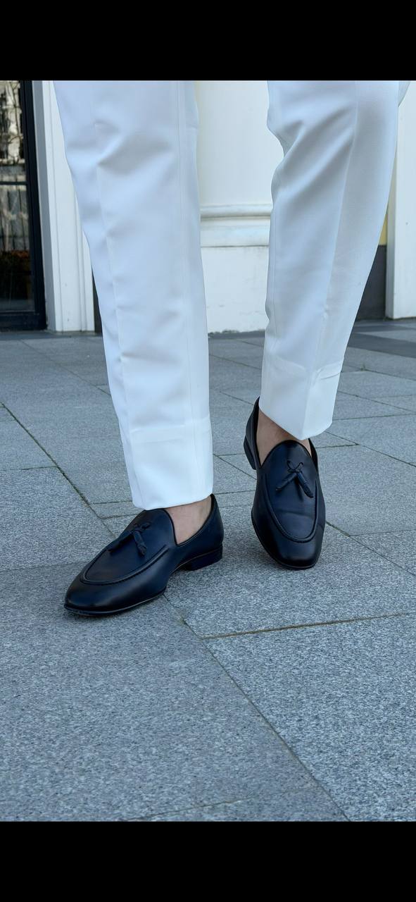 Men’s black leather tassel loafers paired with white tailored trousers, showcasing a sleek slip-on design and classic tassel detail.