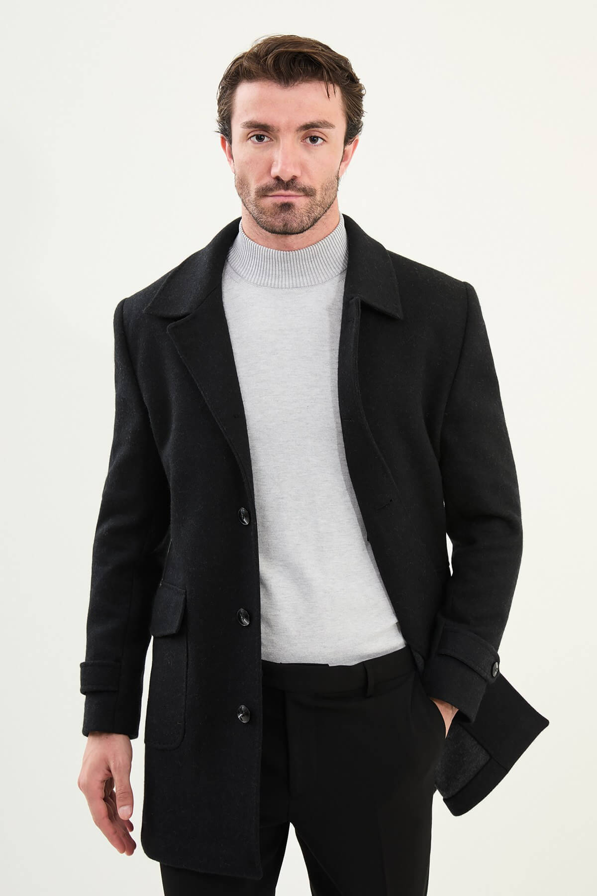 A man wearing a slim fit black single-breasted coat over a light grey turtleneck, styled with black trousers and black leather boots.