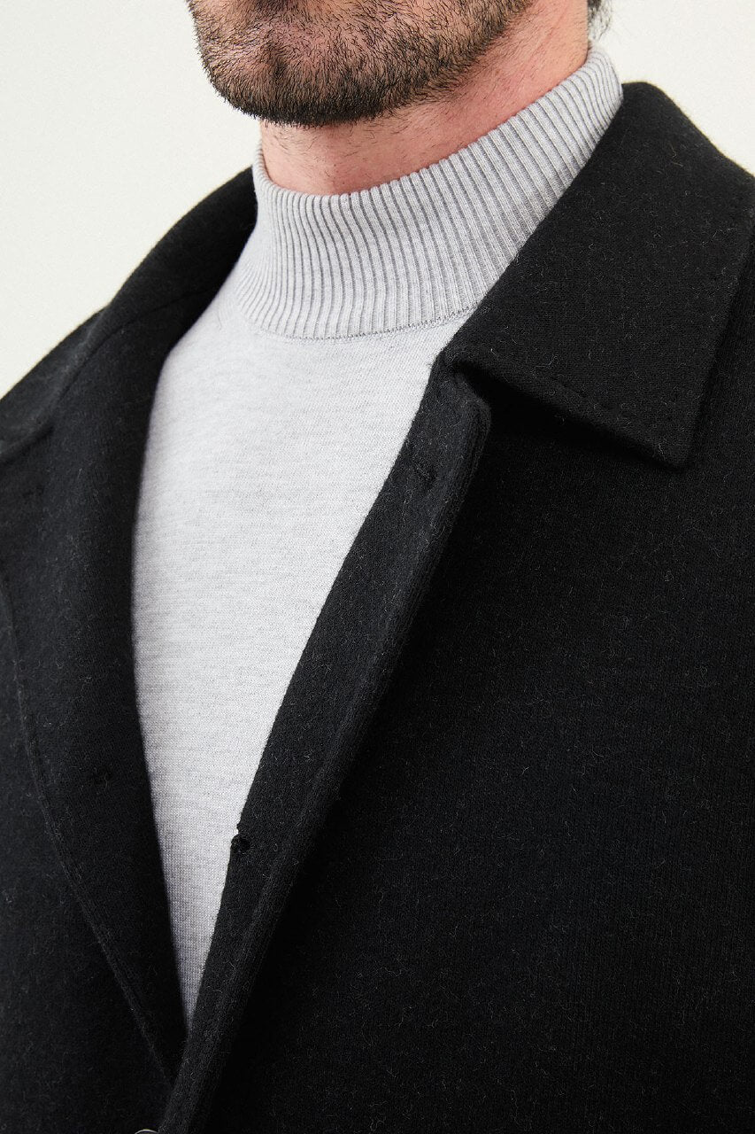 A man wearing a slim fit black single-breasted coat over a light grey turtleneck, styled with black trousers and black leather boots.