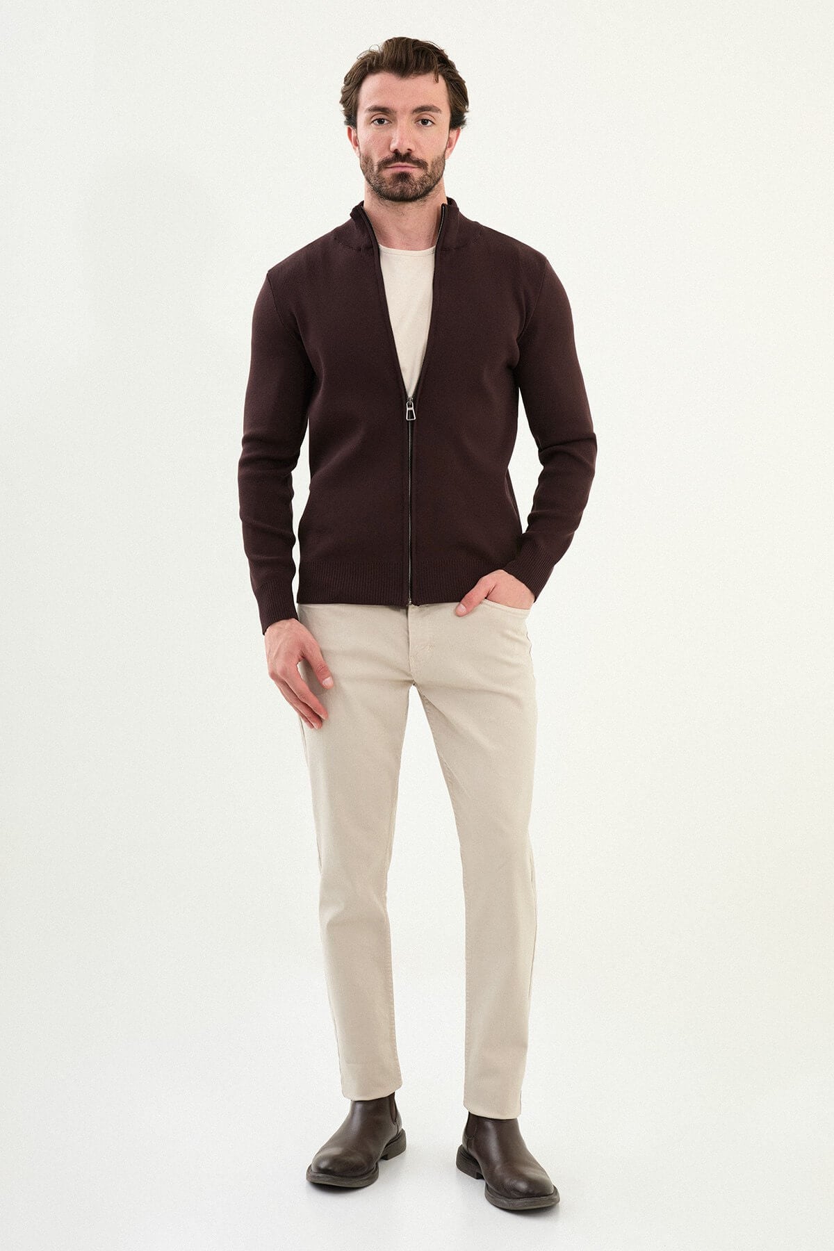 A man wearing a slim fit brown zip cardigan over a beige T-shirt, styled with light beige trousers.