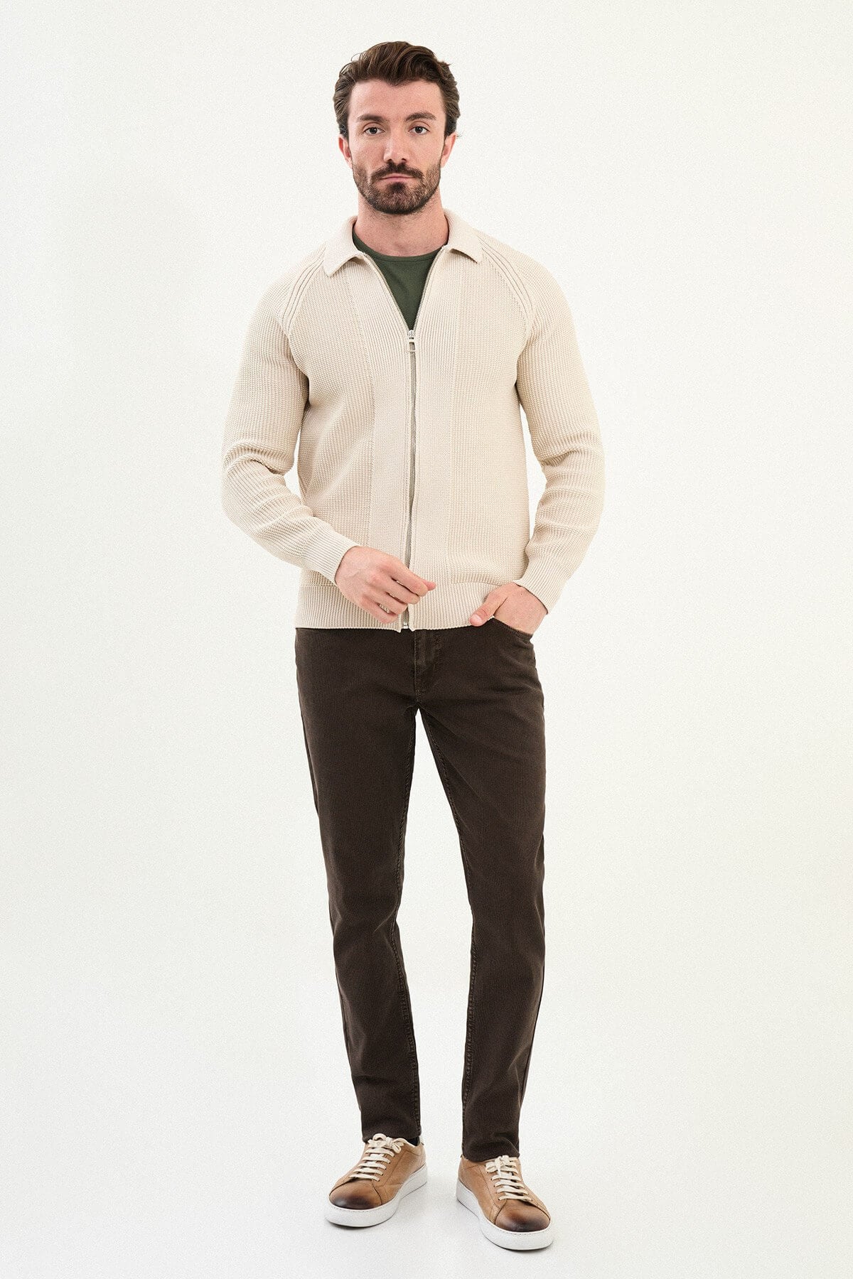 A man wearing a slim fit beige ribbed zip-up cardigan over a green crewneck t-shirt, styled with brown trousers and tan sneakers.