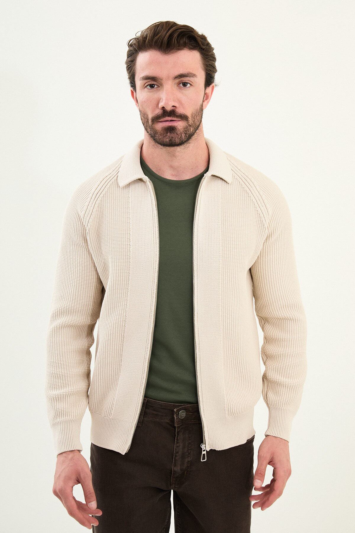 A man wearing a slim fit beige ribbed zip-up cardigan over a green crewneck t-shirt, styled with brown trousers and tan sneakers.