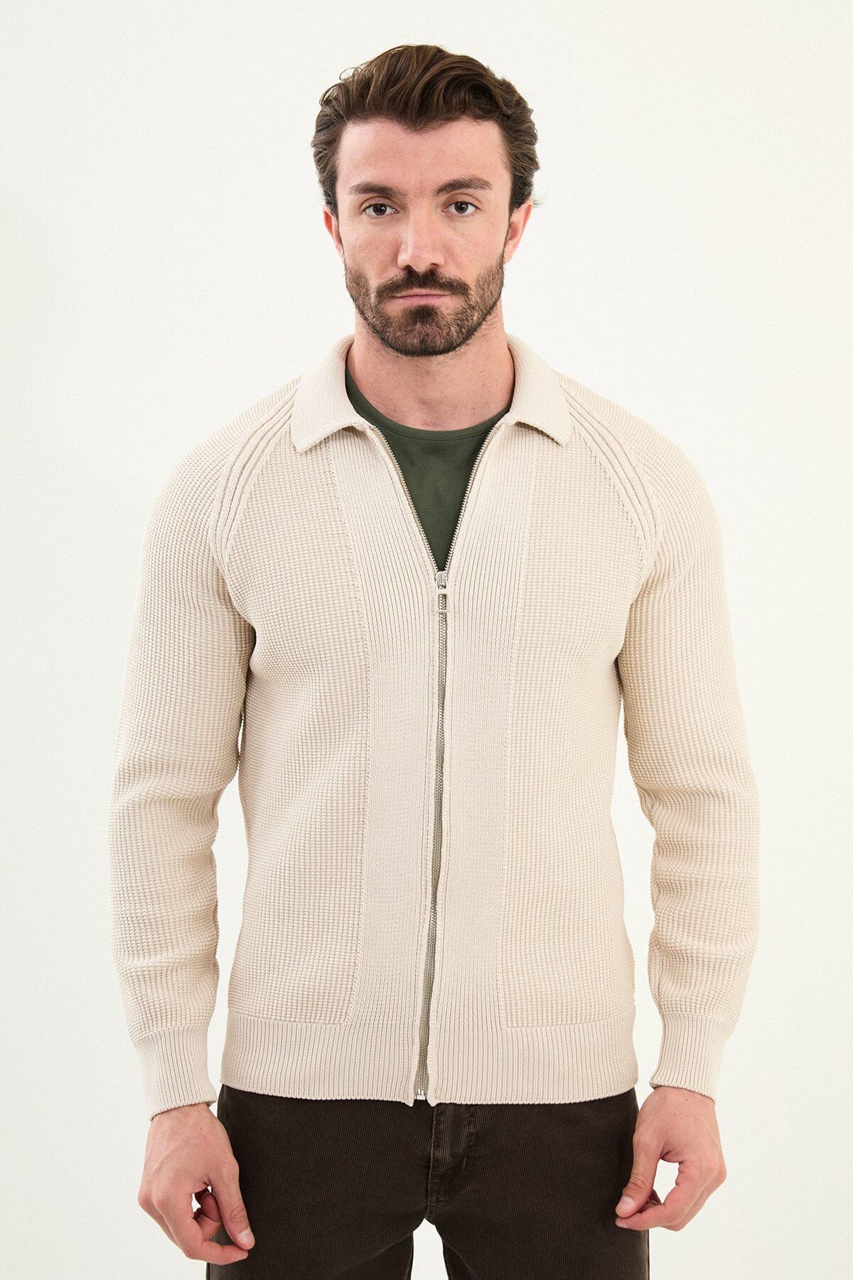 A man wearing a slim fit beige ribbed zip-up cardigan over a green crewneck t-shirt, styled with brown trousers and tan sneakers.